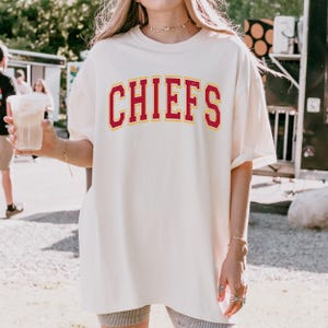May include: Cream-colored t-shirt with the word "CHIEFS" in red block letters with a yellow outline. The shirt is a relaxed fit and appears to be made of a soft material. The shirt is part of a sports apparel collection.