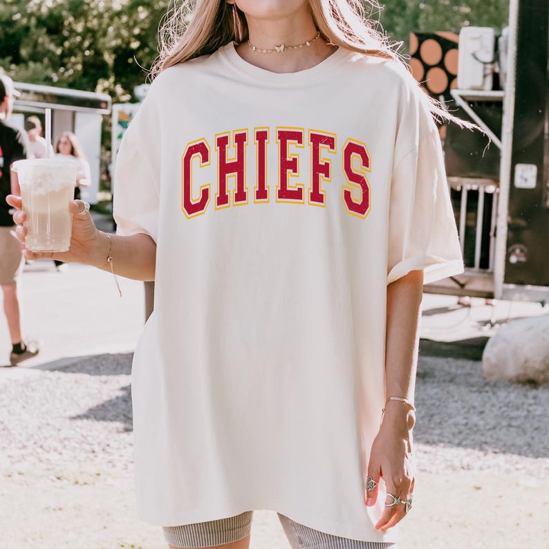 May include: Cream-colored t-shirt with the word "CHIEFS" in red block letters with a yellow outline. The shirt is a relaxed fit and appears to be made of a soft material. The shirt is part of a sports apparel collection.