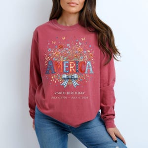 May include: A maroon long-sleeve sweatshirt with a floral and butterfly design. The word "AMERICA" is displayed in red and blue letters. A blue and white plaid bow is below the word. The text "250th BIRTHDAY JULY 4, 1776 - JULY 4, 2026" is printed below the bow.