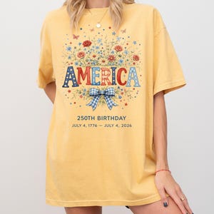 May include: Mustard yellow t-shirt with a floral design and the word "AMERICA" in red, white, and blue. The shirt celebrates the 250th birthday, with the dates July 4, 1776 - July 4, 2026. The design includes a blue and white bow.