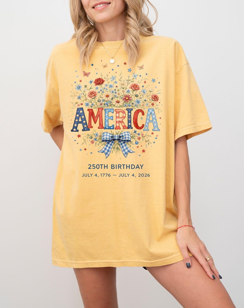 May include: Mustard yellow t-shirt with a floral design and the word "AMERICA" in red, white, and blue. The shirt celebrates the 250th birthday, with the dates July 4, 1776 - July 4, 2026. The design includes a blue and white bow.