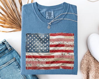 May include: A blue t-shirt with a sewn-on American flag design. The flag features a patchwork design with red and white stripes and a blue field of stars. A silver necklace and blue jeans are also in the image.