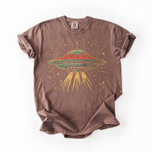 May include: A brown short-sleeve t-shirt featuring a graphic print of a flying saucer emitting light beams downward. The saucer is colored in shades of red, green, and yellow, surrounded by small star shapes. The shirt has a crew neckline.