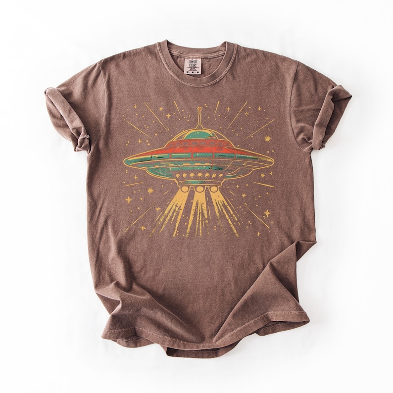 May include: A brown short-sleeve t-shirt featuring a graphic print of a flying saucer emitting light beams downward. The saucer is colored in shades of red, green, and yellow, surrounded by small star shapes. The shirt has a crew neckline.