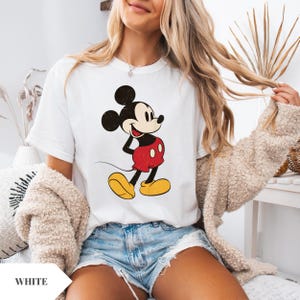 May include: White t-shirt featuring a vintage-style Mickey Mouse graphic. The cartoon character is depicted in his classic red shorts, yellow shoes, and black ears. The word "WHITE" is visible in the bottom left corner.