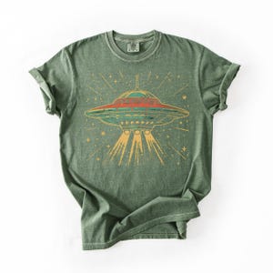 May include: A sage green short-sleeve t-shirt featuring a graphic print of a flying saucer emitting light beams downward. The design includes a red and teal UFO surrounded by yellow stars and radiating lines on the front of the garment.