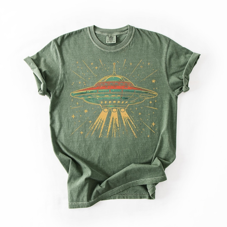 May include: A sage green short-sleeve t-shirt featuring a graphic print of a flying saucer emitting light beams downward. The design includes a red and teal UFO surrounded by yellow stars and radiating lines on the front of the garment.