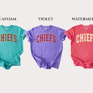 Op de afbeelding: Drie t-shirts met korte mouwen in de kleuren zeegroen, violet en watermeloen. Elk shirt heeft het woord CHIEFS in een gebogen, collegestijl lettertype op de borst gedrukt. De zeegroene en violette shirts hebben rode letters, terwijl het watermeloenkleurige shirt witte letters heeft.