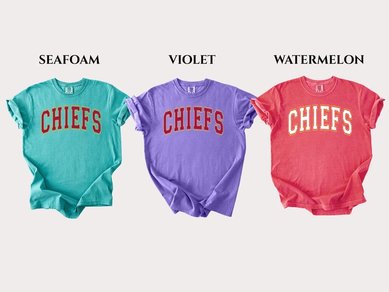 Op de afbeelding: Drie t-shirts met korte mouwen in de kleuren zeegroen, violet en watermeloen. Elk shirt heeft het woord CHIEFS in een gebogen, collegestijl lettertype op de borst gedrukt. De zeegroene en violette shirts hebben rode letters, terwijl het watermeloenkleurige shirt witte letters heeft.
