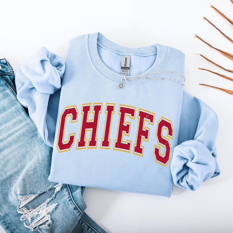 May include: A light blue sweatshirt with the word "CHIEFS" in red letters with a gold outline. The sweatshirt is folded with the sleeves tucked in. A silver necklace with a small pendant is on the sweatshirt. A pair of blue jeans is visible.
