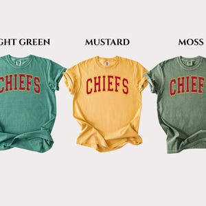 Op de afbeelding: Drie T-shirts met korte mouwen en ronde hals in de kleuren lichtgroen, mosterdgeel en mosgroen. Elk shirt heeft het woord CHIEFS in een rood gebogen lettertype met een gouden omlijning op de borst. De labels LIGHT GREEN, MUSTARD, MOSS staan erboven.