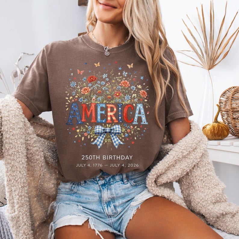 May include: A brown t-shirt with a floral design and the word "AMERICA" in red, white, and blue. The design includes a blue and white bow and the text "250th BIRTHDAY JULY 4, 1776 &ndash; JULY 4, 2026".