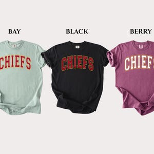Op de afbeelding: Drie t-shirts met korte mouwen in de kleuren bay, zwart en berry. Elk shirt heeft het woord CHIEFS in een gebogen, collegiale lettertype op de borst gedrukt. Het bay shirt heeft rode tekst, het zwarte shirt heeft rode tekst en het berry shirt heeft witte tekst.