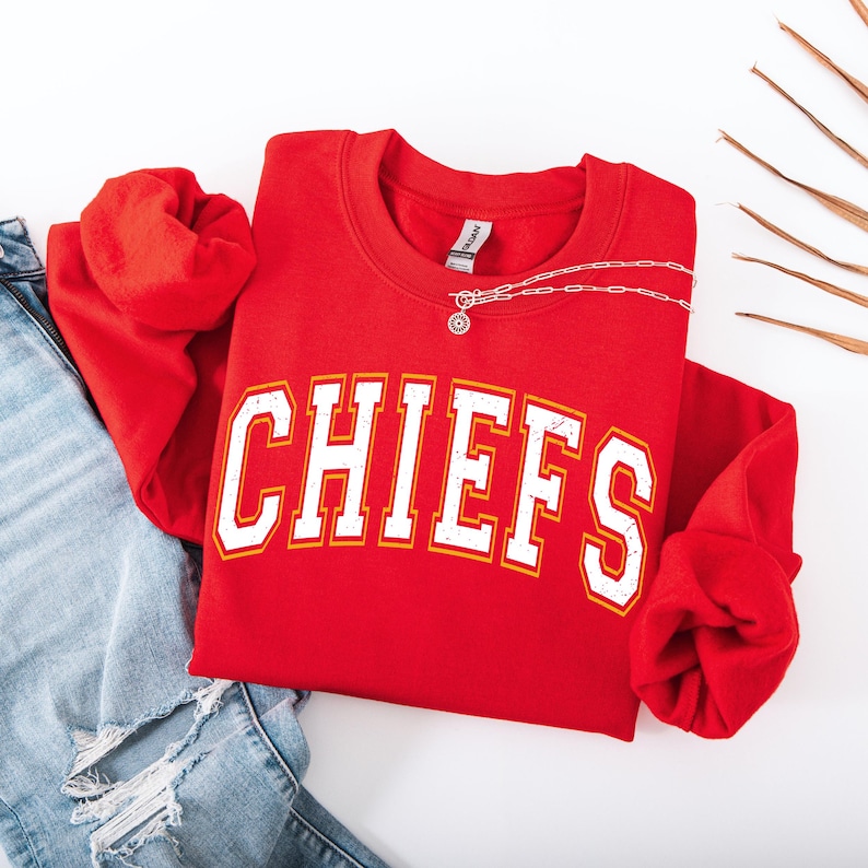 K&ouml;nnte beinhalten: Ein rotes Sweatshirt mit Rundhalsausschnitt und dem Wort "CHIEFS" in wei&szlig;en Blockbuchstaben mit gelbem Rand. Eine silberne Halskette mit Anh&auml;nger liegt &uuml;ber dem Sweatshirt. Das Sweatshirt wird mit einer blauen Jeans kombiniert.