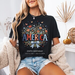 May include: Black t-shirt with a floral design and the word "AMERICA" in red, white, and blue. The shirt also features the text "250TH BIRTHDAY JULY 4, 1776 &ndash; JULY 4, 2026".