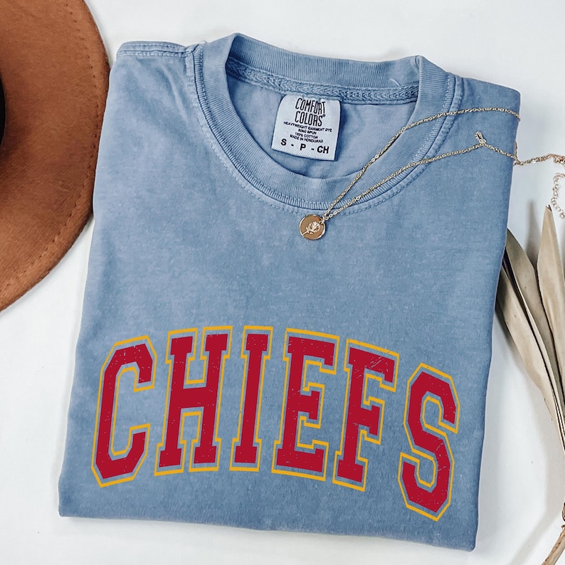 May include: A light blue t-shirt with the word "CHIEFS" in red letters with a yellow outline. A gold necklace with a pendant is draped across the shirt. The shirt is folded on a white surface.