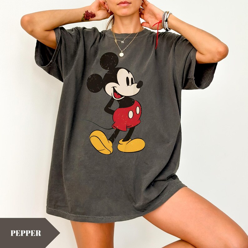 May include: Dark gray t-shirt featuring a distressed graphic of Mickey Mouse in his classic pose. The cartoon character is wearing red shorts, yellow shoes, and a white-gloved hand is on his hip. The shirt has short sleeves and a relaxed fit.