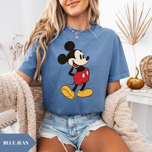 May include: A blue jean t-shirt featuring a classic Mickey Mouse graphic. The cartoon character is depicted in his iconic red shorts, yellow shoes, and black gloves. The shirt has short sleeves and a relaxed fit, perfect for casual wear.