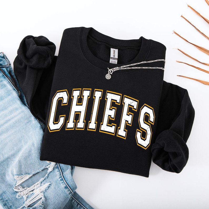 May include: Black sweatshirt with the word "CHIEFS" in white, outlined in gold. A silver necklace with a pendant is draped across the sweatshirt. The sweatshirt is paired with blue jeans.