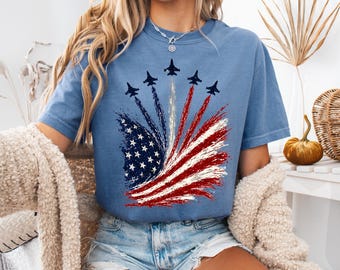 May include: A blue short-sleeve t-shirt featuring a graphic print of five fighter jets flying in formation, leaving red, white, and blue smoke trails that form a stylized American flag design with stars and stripes on the front of the garment.