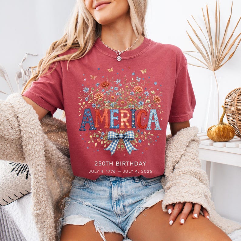May include: A maroon t-shirt with the word "AMERICA" in a floral design. The shirt has a floral arrangement above the word and a blue and white bow below. The text "250TH BIRTHDAY JULY 4, 1776 - JULY 4, 2026" is printed below the bow.