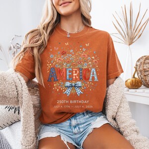 May include: Burnt orange t-shirt with the word "AMERICA" in a floral design. The shirt features a blue and white gingham bow and the text "250TH BIRTHDAY JULY 4, 1776 - JULY 4, 2026."