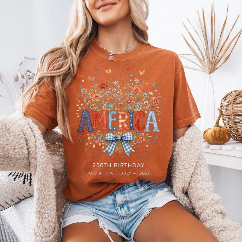May include: Burnt orange t-shirt with the word "AMERICA" in a floral design. The shirt features a blue and white gingham bow and the text "250TH BIRTHDAY JULY 4, 1776 - JULY 4, 2026."