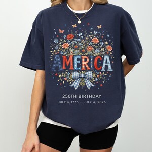 May include: Navy blue t-shirt with a floral design and the word "AMERICA" in red, white, and blue letters. The shirt commemorates the 250th birthday, with the dates July 4, 1776 - July 4, 2026.