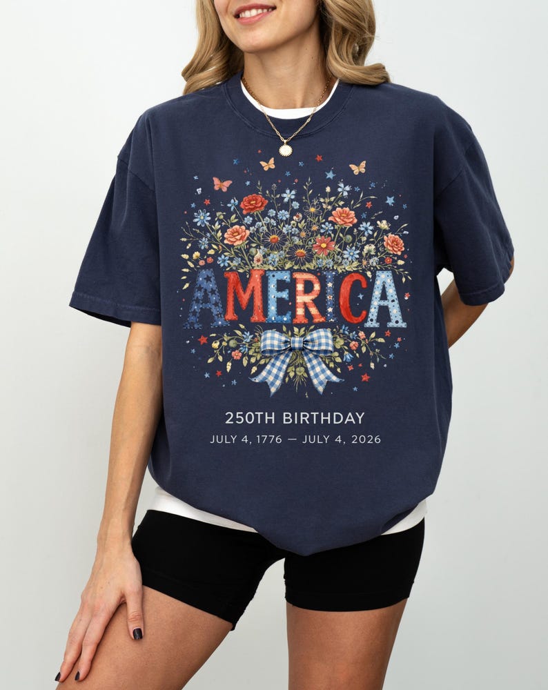 May include: Navy blue t-shirt with a floral design and the word "AMERICA" in red, white, and blue letters. The shirt commemorates the 250th birthday, with the dates July 4, 1776 - July 4, 2026.