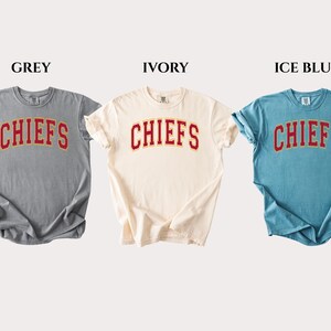 Op de afbeelding: Drie T-shirts met korte mouwen en ronde hals in grijs, ivoor en ijsblauw. Elk shirt heeft het woord CHIEFS in een rood gebogen lettertype met een gele omlijning op de borst. De shirts worden naast elkaar getoond met labels erboven.
