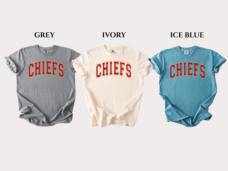 Op de afbeelding: Drie T-shirts met korte mouwen en ronde hals in grijs, ivoor en ijsblauw. Elk shirt heeft het woord CHIEFS in een rood gebogen lettertype met een gele omlijning op de borst. De shirts worden naast elkaar getoond met labels erboven.