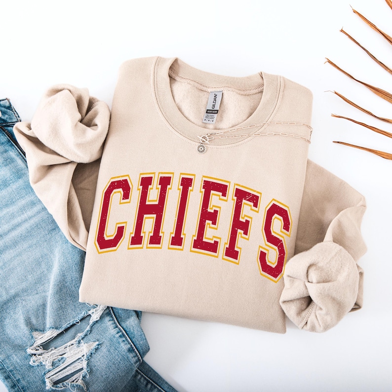 May include: A beige sweatshirt with the word "CHIEFS" in red letters with a gold outline. The sweatshirt is folded and placed on top of a pair of blue jeans. A silver necklace with a small pendant is on the sweatshirt.