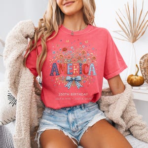 May include: A coral-colored t-shirt with the word "AMERICA" in a floral design. The shirt features a blue and white gingham bow and the text "250TH BIRTHDAY JULY 4, 1776 - JULY 4, 2026". The shirt is worn with denim shorts.