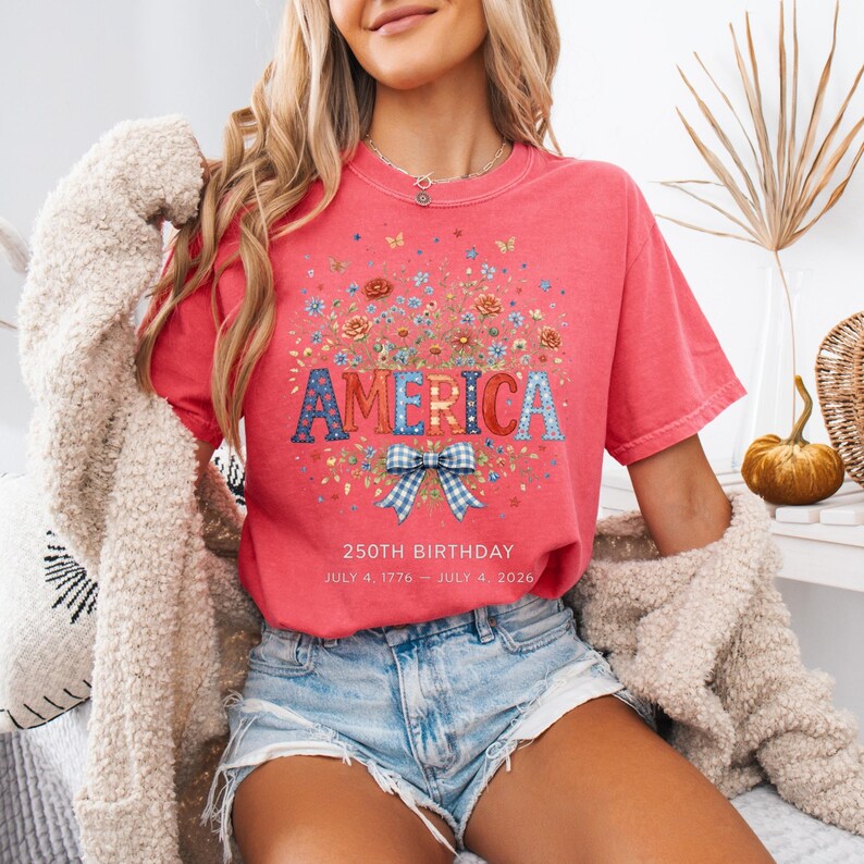 May include: A coral-colored t-shirt with the word "AMERICA" in a floral design. The shirt features a blue and white gingham bow and the text "250TH BIRTHDAY JULY 4, 1776 - JULY 4, 2026". The shirt is worn with denim shorts.