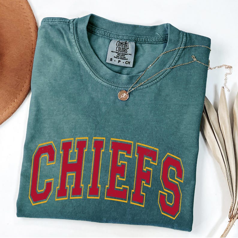 May include: A teal Comfort Colors t-shirt with the word "CHIEFS" in red letters with a yellow outline. A gold necklace with a pendant rests on the shirt. The shirt is folded and has a small tag at the neckline.