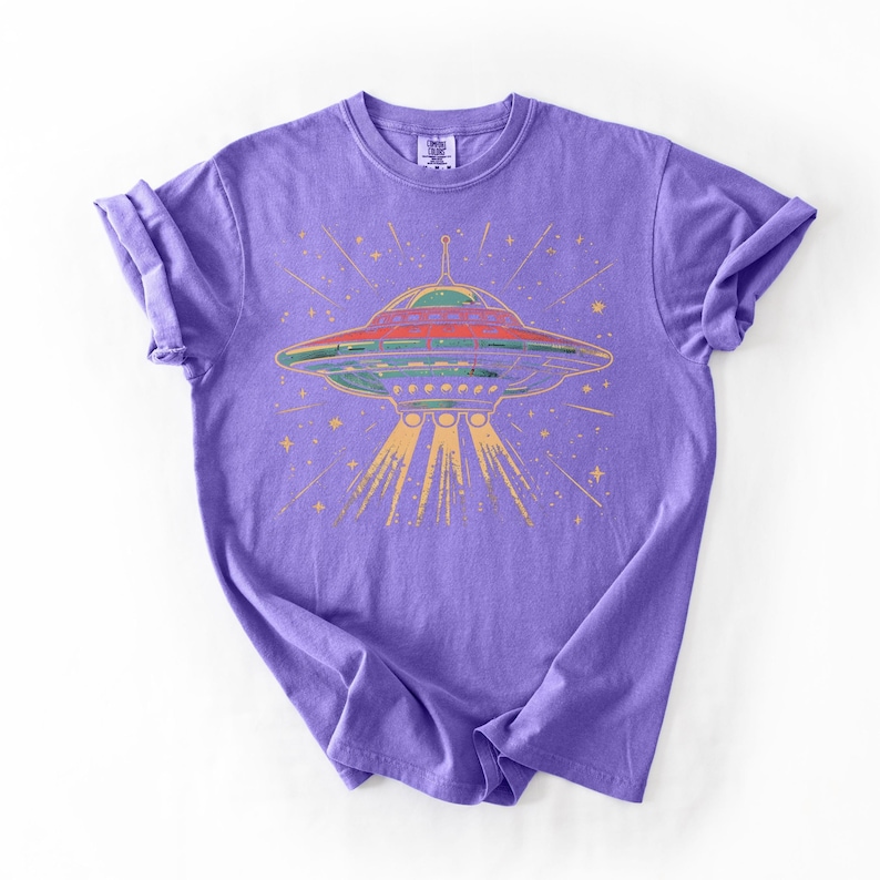 May include: A purple short-sleeve t-shirt featuring a graphic print of a flying saucer emitting yellow light beams. The design includes small yellow stars surrounding the spacecraft, which is rendered in shades of green, red, and yellow.