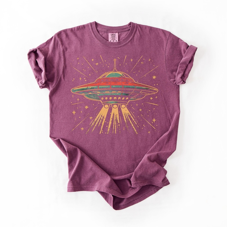 May include: A plum-colored short-sleeve t-shirt featuring a graphic print of a flying saucer emitting yellow light beams. The design includes a star-filled background and a small label at the inner neckline. The fabric has a slightly textured appearance.