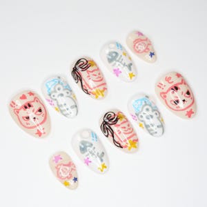 May include: A collection of ten almond-shaped false nails. Each nail features a unique design, including cartoon cats, fish bones, bows, and stars. The colour scheme includes pink, black, white, and yellow on a nude base.