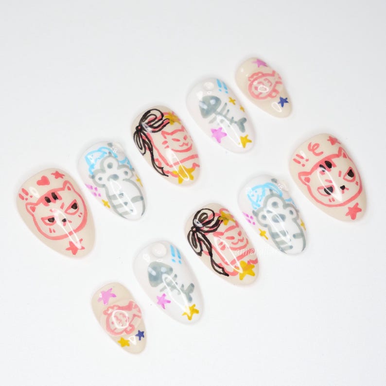 May include: A collection of ten almond-shaped false nails. Each nail features a unique design, including cartoon cats, fish bones, bows, and stars. The colour scheme includes pink, black, white, and yellow on a nude base.