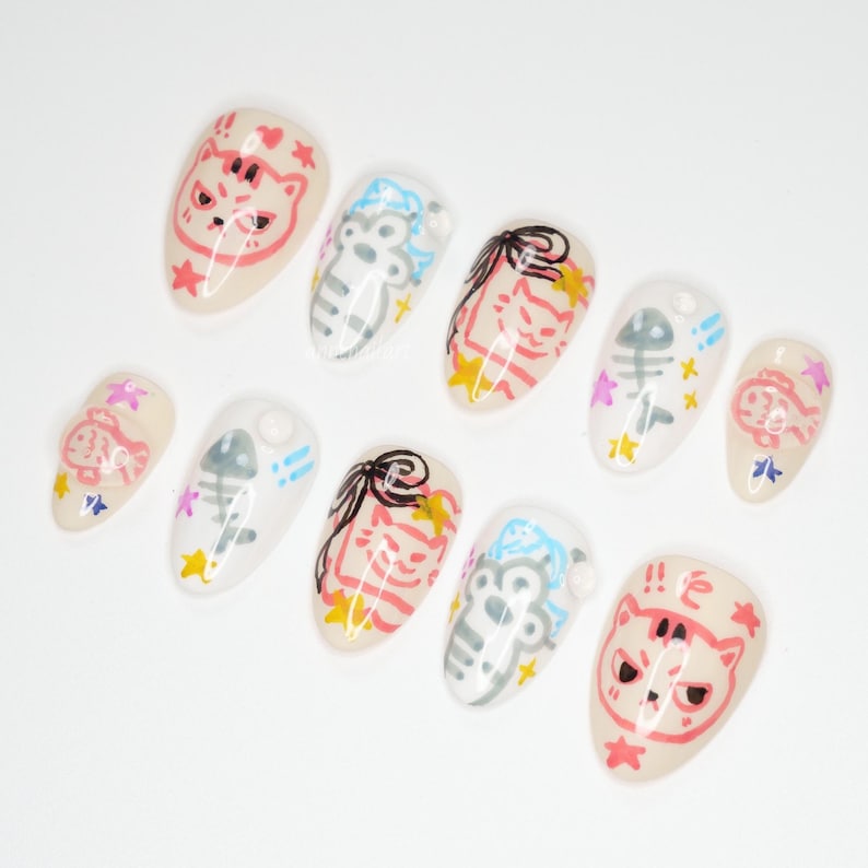 May include: A collection of ten artificial nails, each adorned with playful cat and fish designs. The nails showcase a palette of pink, black, white, and blue, complemented by star and fishbone motifs. The oval-shaped nails have a glossy finish.