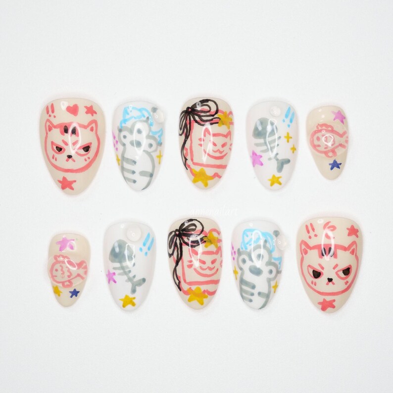 May include: A set of ten almond-shaped false nails. Each nail features a unique design, including cats, fish bones, bows, and stars. The colour palette includes beige, pink, white, grey, and yellow.