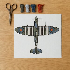 May include: A cross-stitch design of a military airplane in shades of green, blue, and brown, with orange and white accents. The airplane is centered on a white canvas, with scissors and spools of thread in various colors arranged around it.