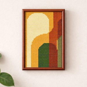 May include: A framed cross-stitch artwork featuring an abstract design in shades of yellow, orange, green, and brown. The artwork is displayed in a dark brown wooden frame and mounted on a white wall. The design has a retro, geometric aesthetic.