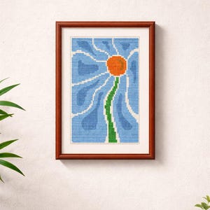 May include: A framed cross-stitch art piece featuring a stylized blue flower with an orange center and green stem. The artwork is set against a light blue background and framed in a dark brown wooden frame. The piece is mounted on a white wall.