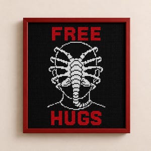 May include: A framed cross-stitch embroidery featuring a black background with a white stitched graphic of a face-hugging creature. The text FREE is stitched in red above the graphic, and the text HUGS is stitched in red below the graphic.