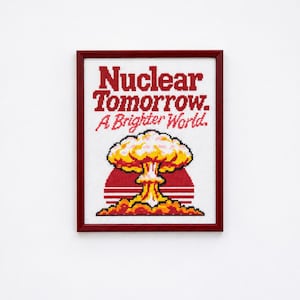 May include: A framed cross-stitch embroidery featuring a red mushroom cloud graphic over a red sun, with the text "Nuclear Tomorrow. A Brighter World." in red lettering on a white fabric background, enclosed in a dark red wooden frame.