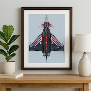May include: Framed artwork featuring a detailed cross-stitch design of a jet fighter. The aircraft is primarily black with red and white accents, and the wings display the Union Jack. The background is a light blue grid.