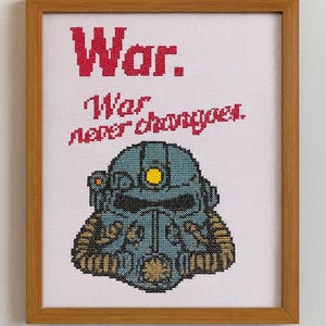 Könnte beinhalten: Kreuzstich-Kunstwerk in einem Holzrahmen. Das Design zeigt einen blau-beigen Helm mit dem Text "War. War never changes." in Rot. Der Hintergrund ist weiß.