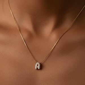 May include: A gold necklace featuring a pendant shaped like the letter "A". The pendant is adorned with small, clear gemstones. The necklace is shown against a plain backdrop.