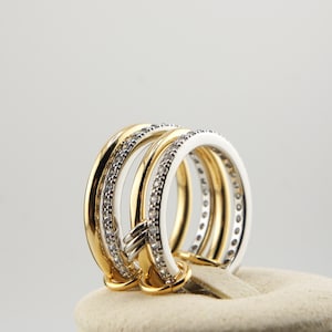 Puede incluir: Un anillo de múltiples bandas con tonos dorados y plateados, adornado con pequeñas piedras brillantes. El anillo se exhibe sobre una superficie de tono neutro, mostrando su intrincado diseño y su elegante atractivo. Las bandas están entrelazadas, creando una pieza de joyería única y llamativa.